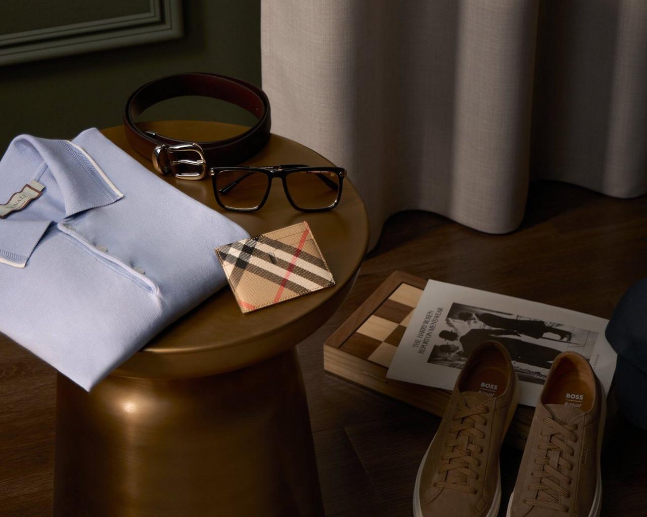 men's accessories, including a light blue collared shirt, a leather belt, eyeglasses, a patterned wallet, and a pair of shoes