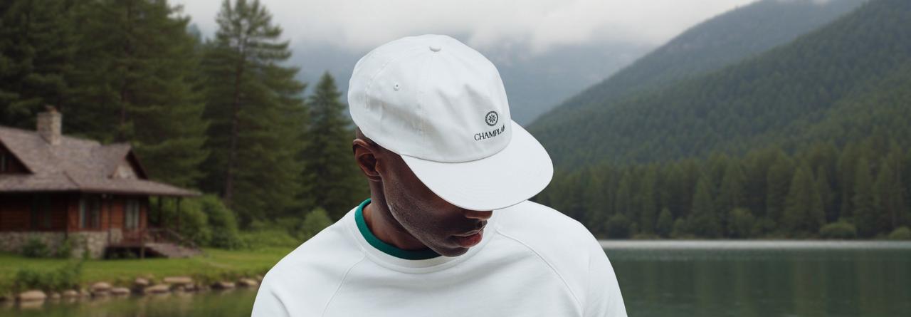  person wearing a white Champlu cap with a small embroidered logo, standing by a calm lake surrounded by forested mountains