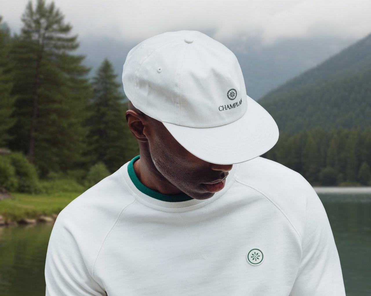  person wearing a white Champlu cap with a small embroidered logo, standing by a calm lake surrounded by forested mountains