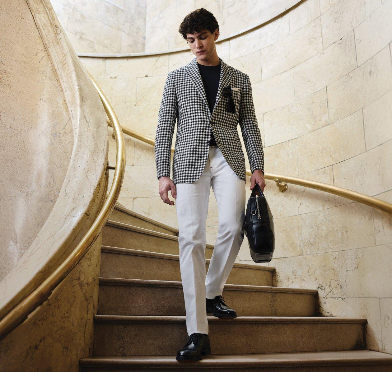 A man wearing a grey and black check patterned coat with black t-shirt and white pants