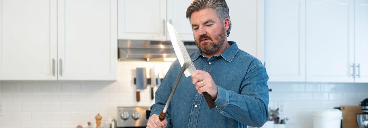 A man in a kitchen holding a large chef’s knife and honing rod while preparing food