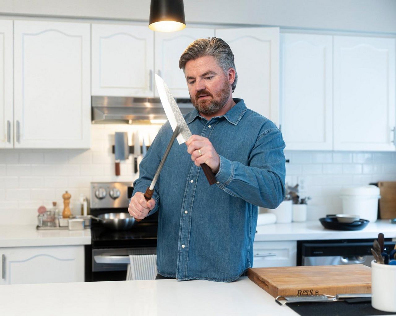 A man in a kitchen holding a large chef’s knife and honing rod while preparing food