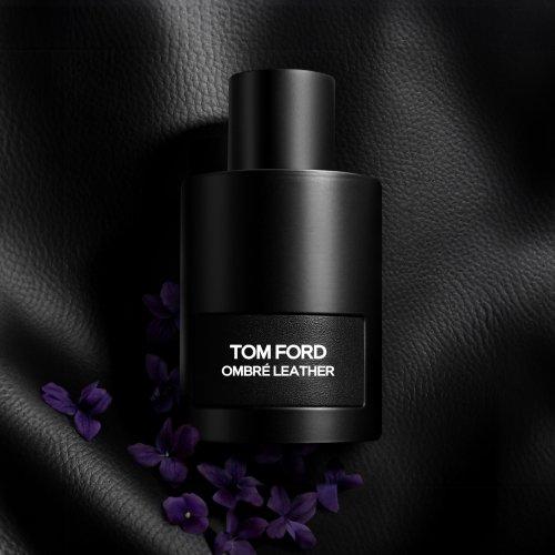 Une bouteille noire de parfum Tom Ford Ombré Leather sur une surface en cuir noir texturé, entourée de petites fleurs violettes