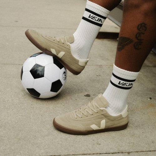Modèle en baskets VEJA Volley en daim beige avec des chaussettes blanches à logo, debout à côté d’un ballon de football classique noir et blanc