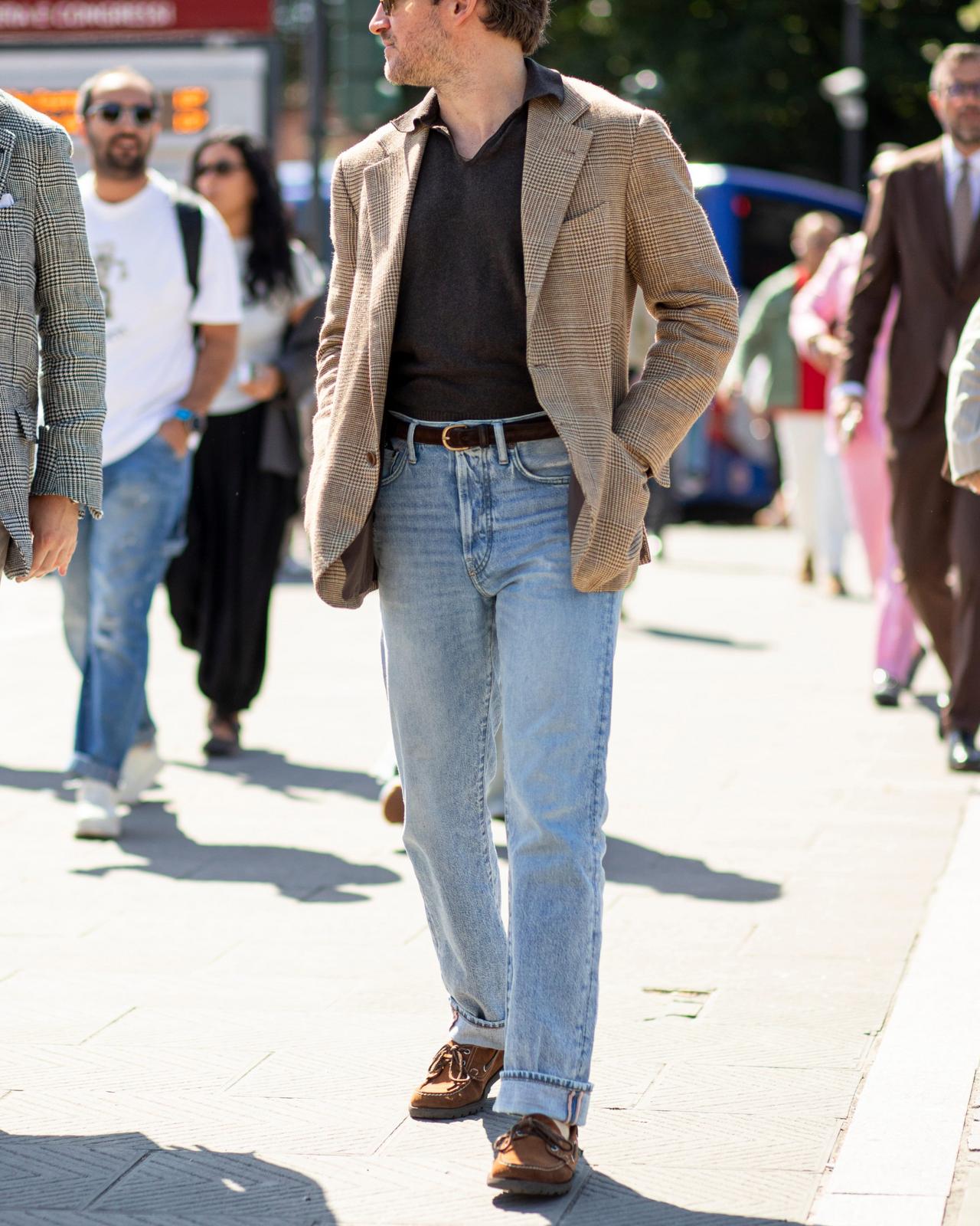 A man featuring a beige plaid blazer, dark V-neck sweater, light blue jeans, and brown leather shoes