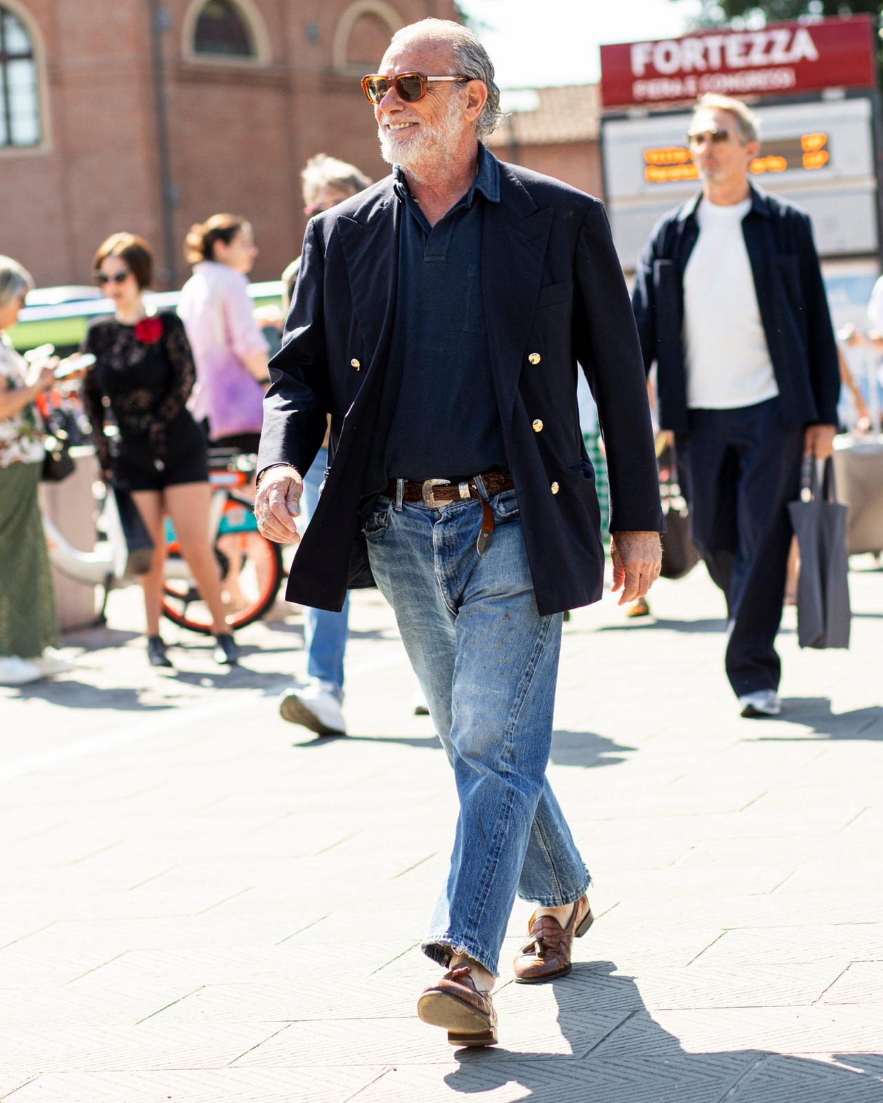 A man wearing a navy double-breasted blazer, dark polo shirt, and light blue jeans