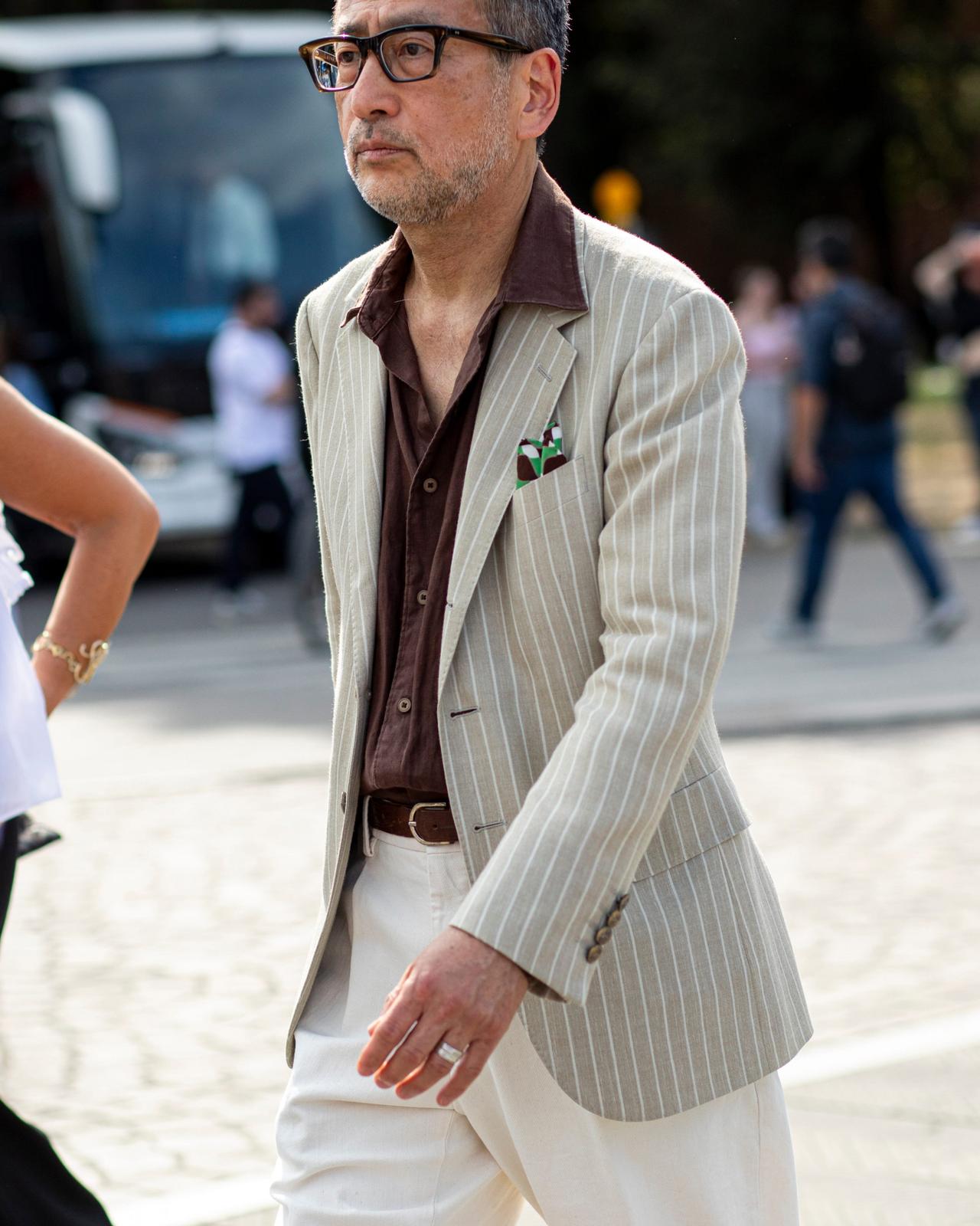 Un homme portant un veston beige pâle à fines rayures par-dessus une chemise brune, agencé à un pantalon blanc et une ceinture assortie