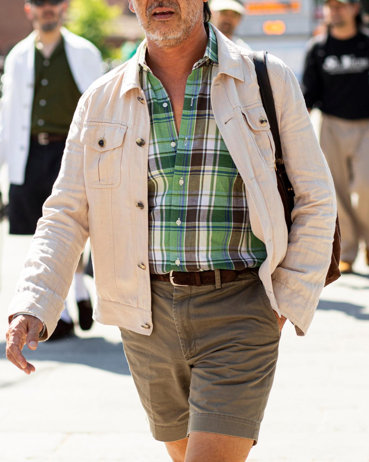 A man walking outdoors wearing a green plaid button-up shirt layered under a light beige jacket, paired with khaki cargo shorts and a brown belt
