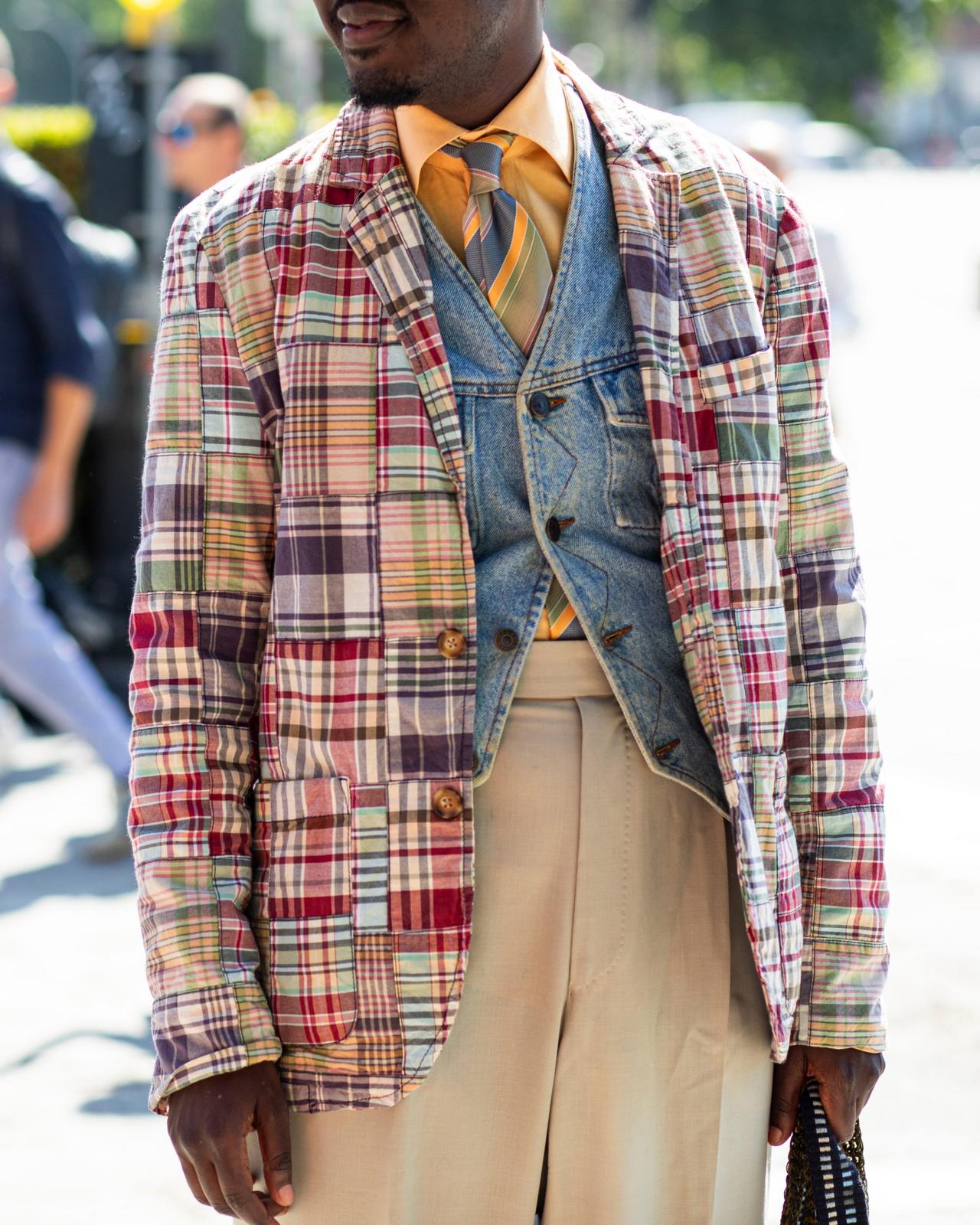 Un homme portant un veston patchwork coloré par-dessus une veste en denim, une chemise jaune et une cravate à rayures, agencé à un pantalon beige