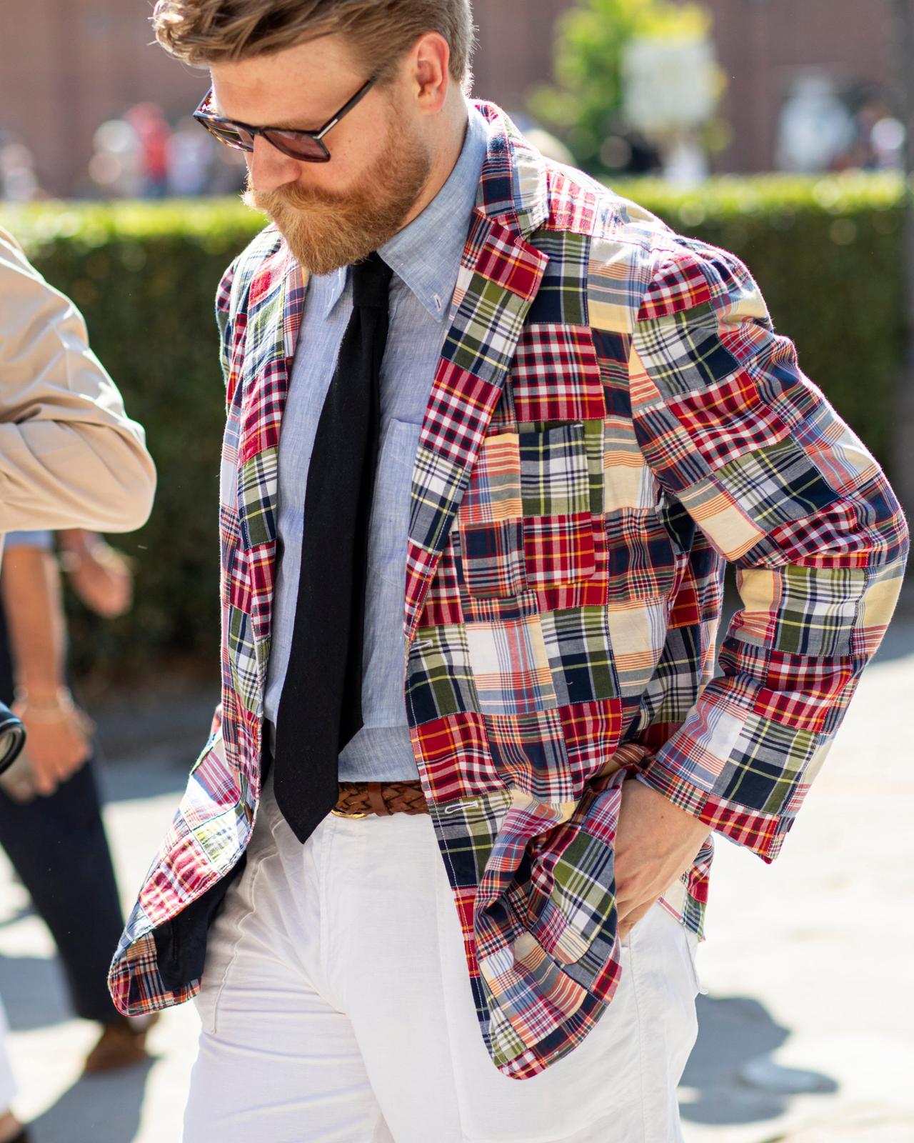 A man wearing a colorful plaid patchwork blazer layered over a denim vest, yellow shirt, and tie, paired with beige trousers