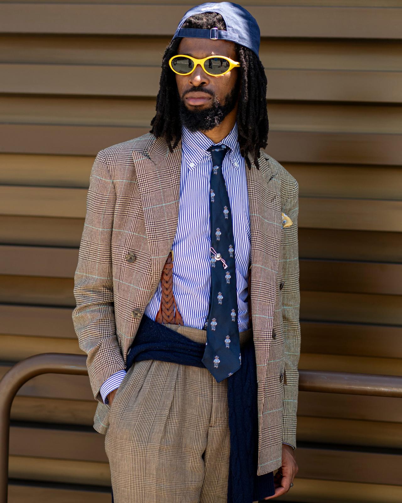 A man wearing brown plaid blazer, striped shirt, and patterned tie layered over a dark sweater