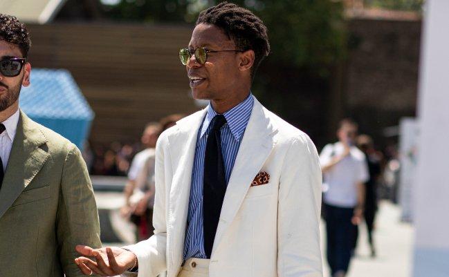 Model in a white suit, blue striped shirt, and dark tie with a pocket square at Pitti Uomo