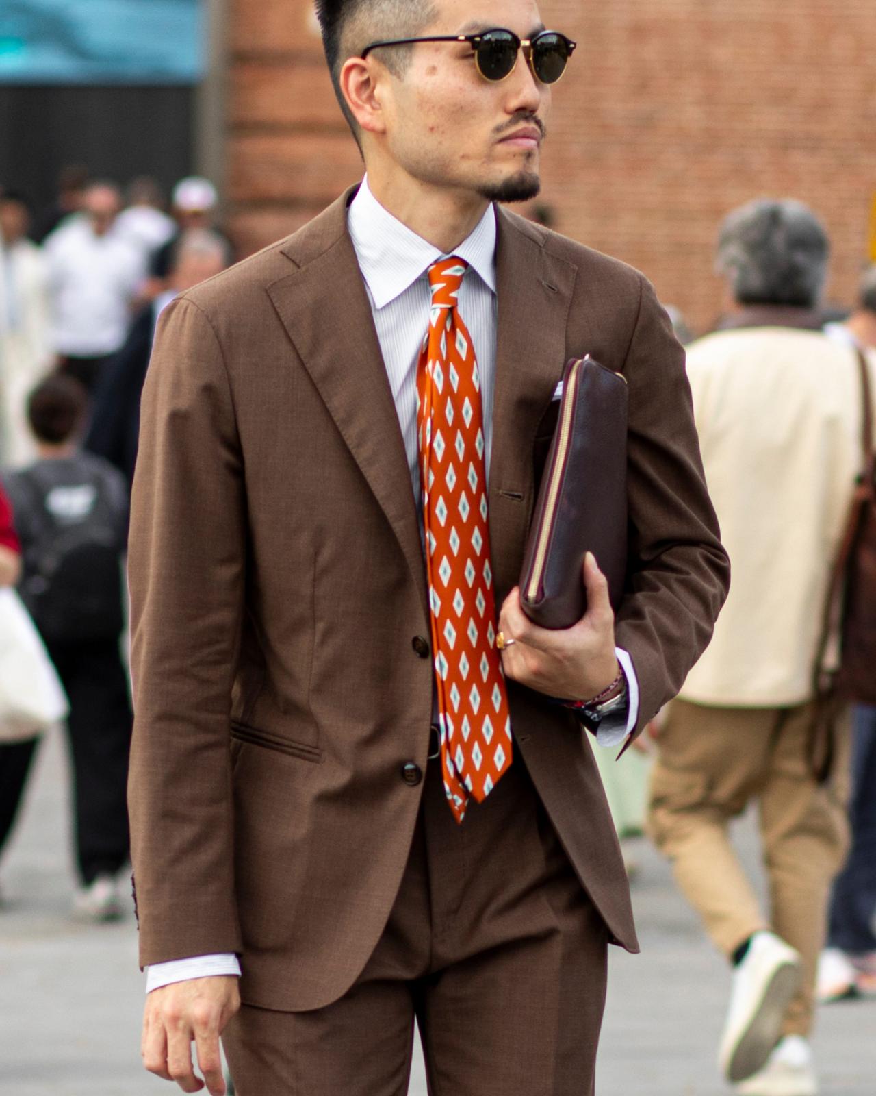 A man in a brown suit paired with a white dress shirt and a bright orange patterned tie, holding a folder in one hand