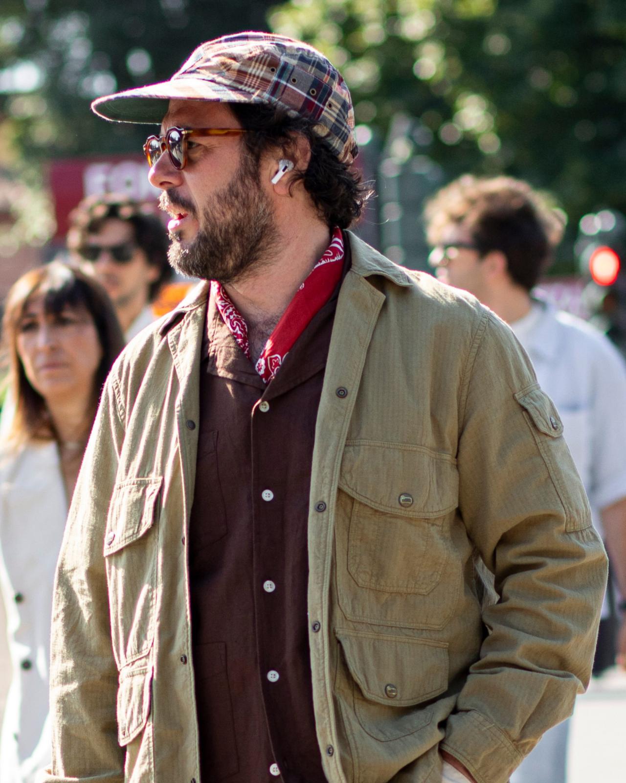 A man wearing a beige field jacket layered over a dark shirt, accessorized with a red bandana and a plaid cap