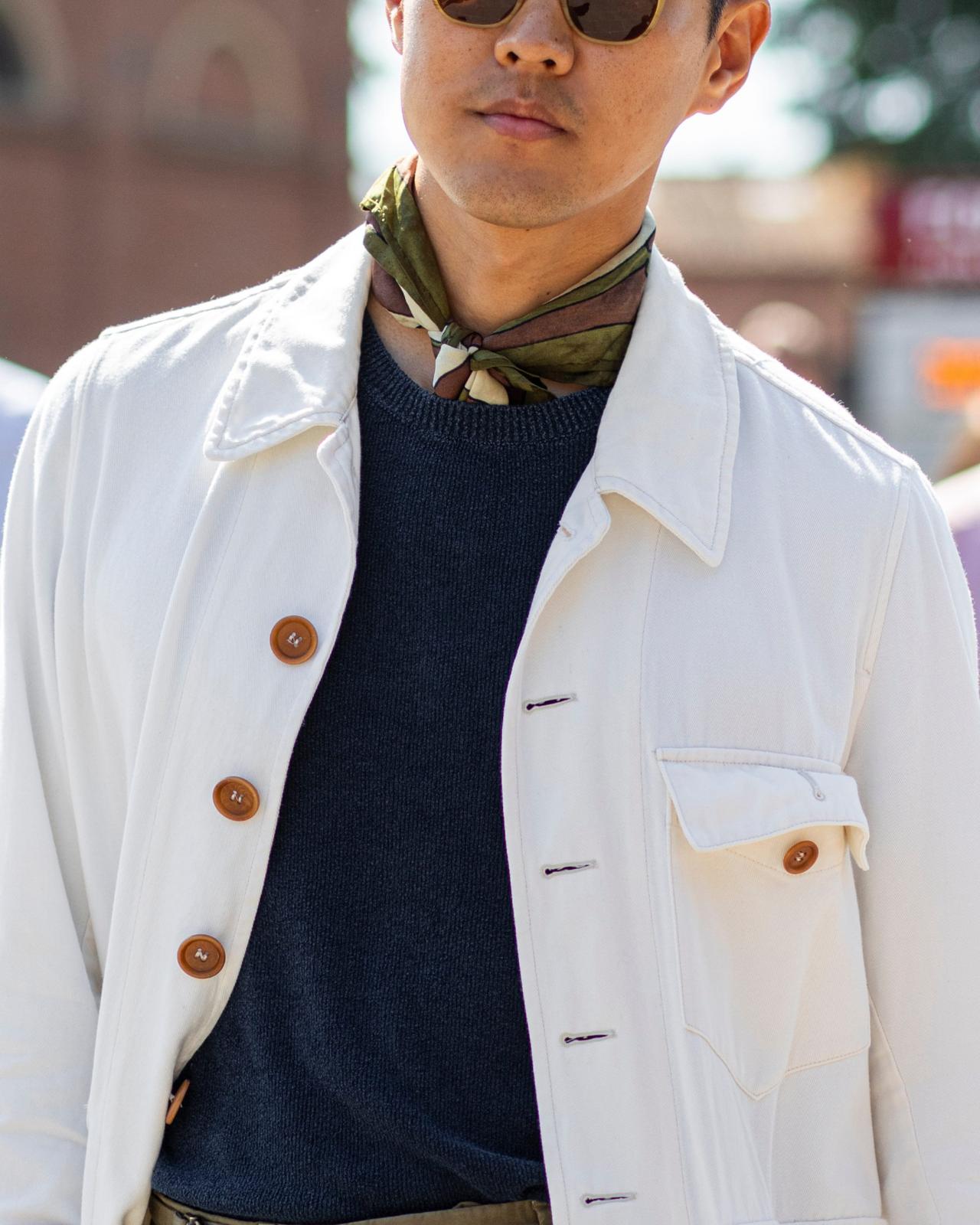 Un homme portant une veste blanche à boutons bruns, accompagnée d’un chandail bleu marine foncé et d’une écharpe à motifs nouée autour du cou
