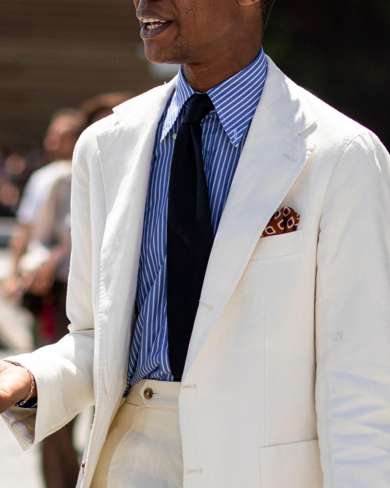 A man wearing a white blazer paired with a blue and white striped shirt, a black tie, and a patterned pocket square