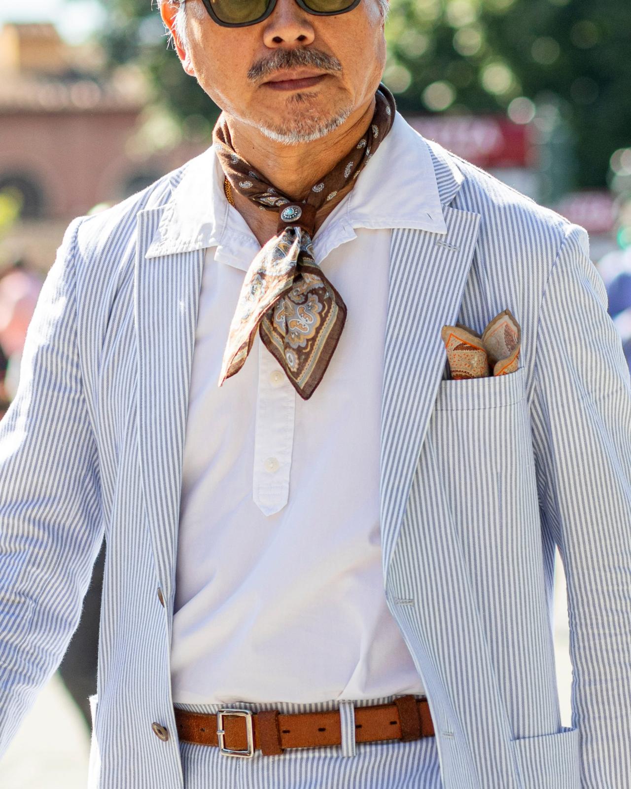 A man wearing a light blue pinstriped suit paired with a white collared shirt, a brown leather belt, and a patterned neck scarf