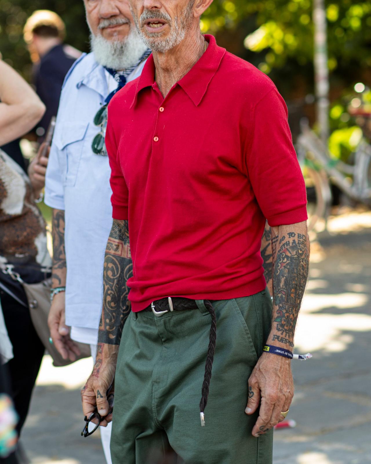 A man wearing a bright red polo shirt paired with olive green trousers, accessorized with a braided belt and visible tattoos on both arms