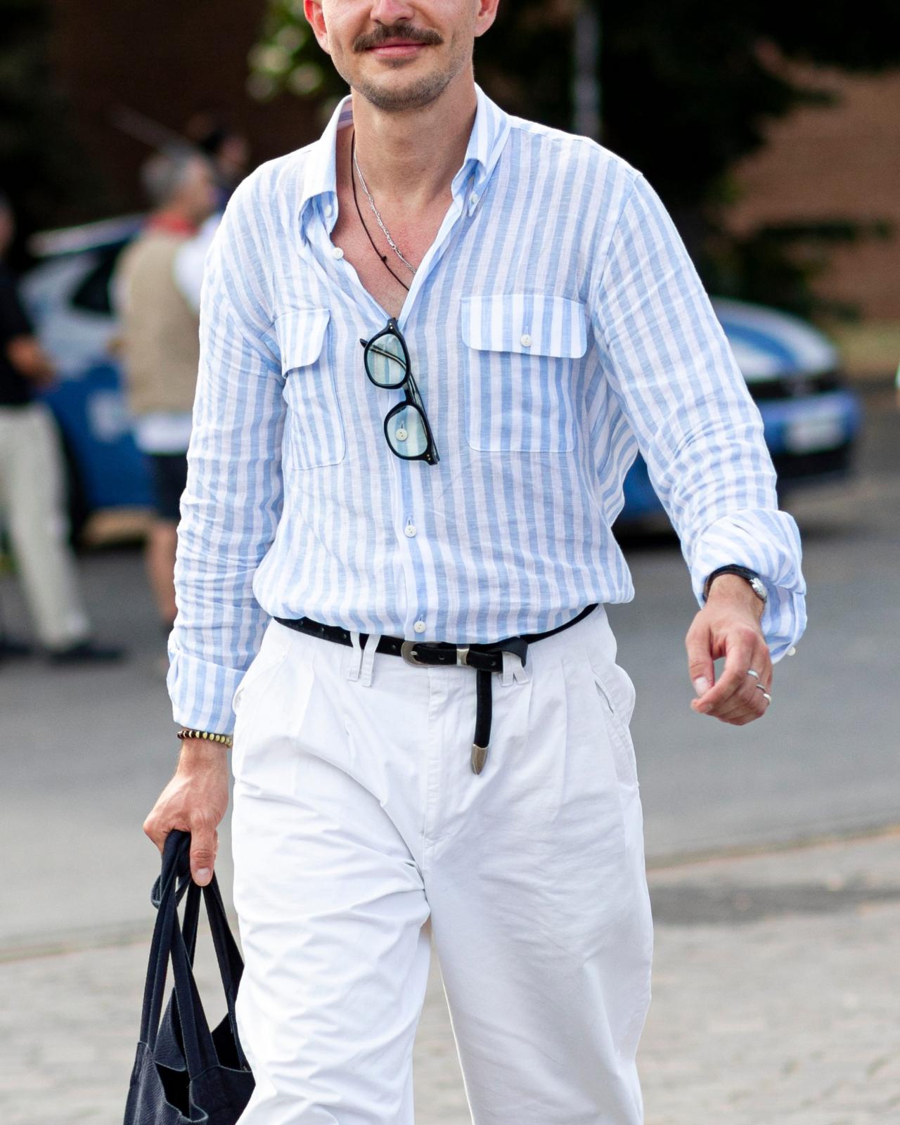 A man wearing a light blue and white striped button-down shirt paired with white trousers and a black belt.