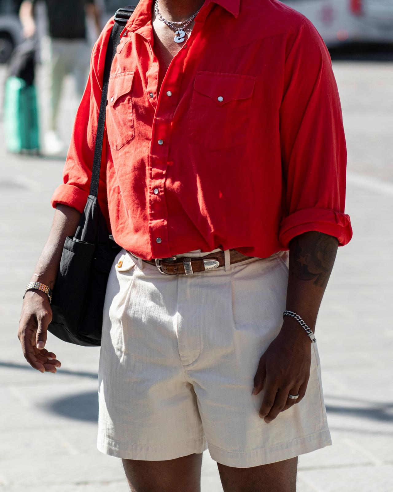 a man wearing a bright red button-up shirt with rolled sleeves, paired with cream-colored pleated shorts and accessorized with a black crossbody bag