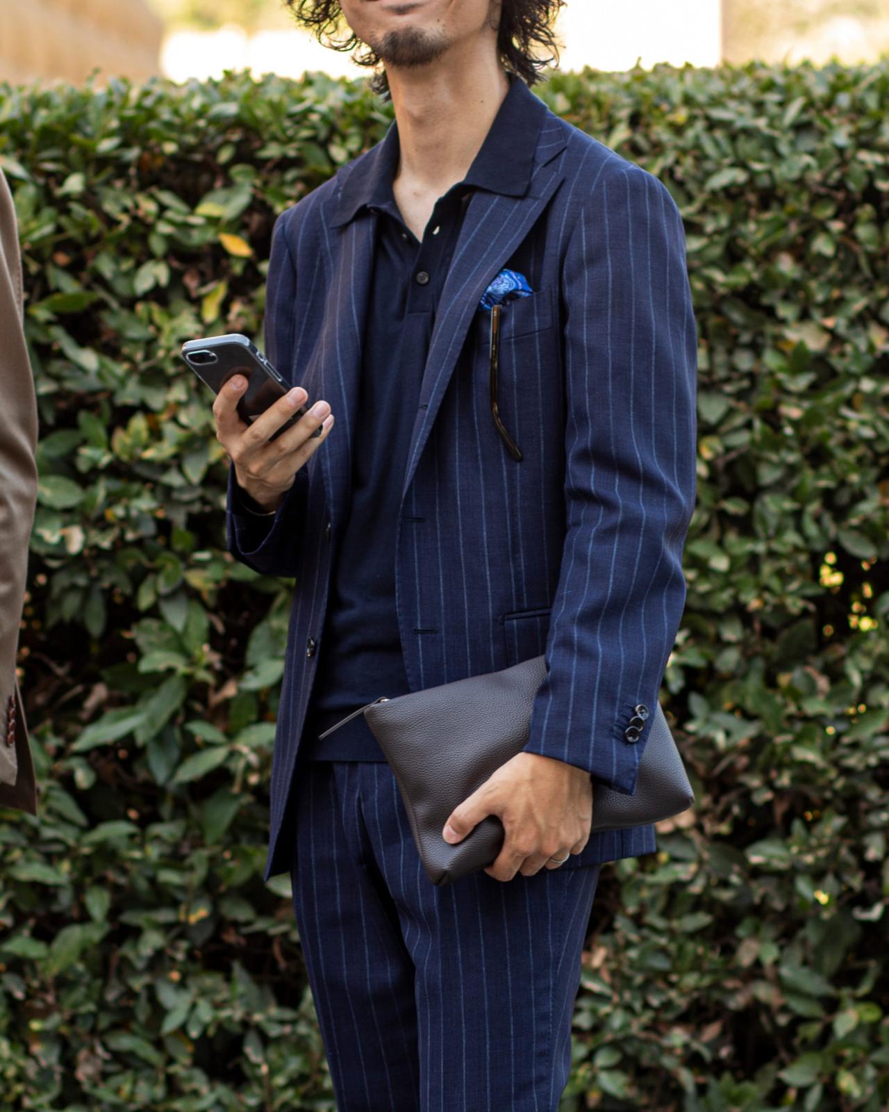Un homme portant un complet bleu marine à fines rayures, accompagné d’un polo foncé, tenant un téléphone intelligent d’une main et une pochette noire de l’autre