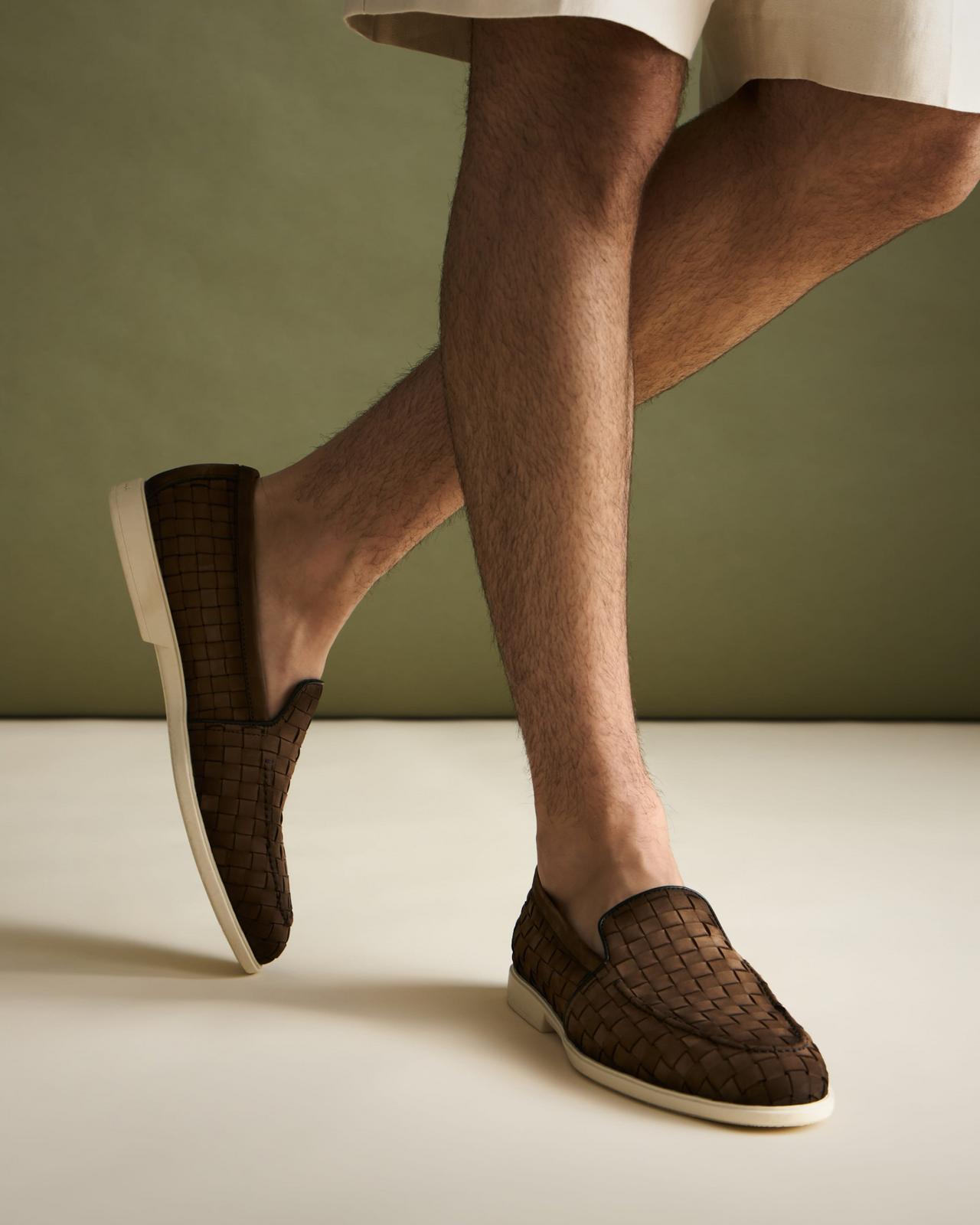 A close-up of a man wearing brown woven slip-on loafers with a white sole