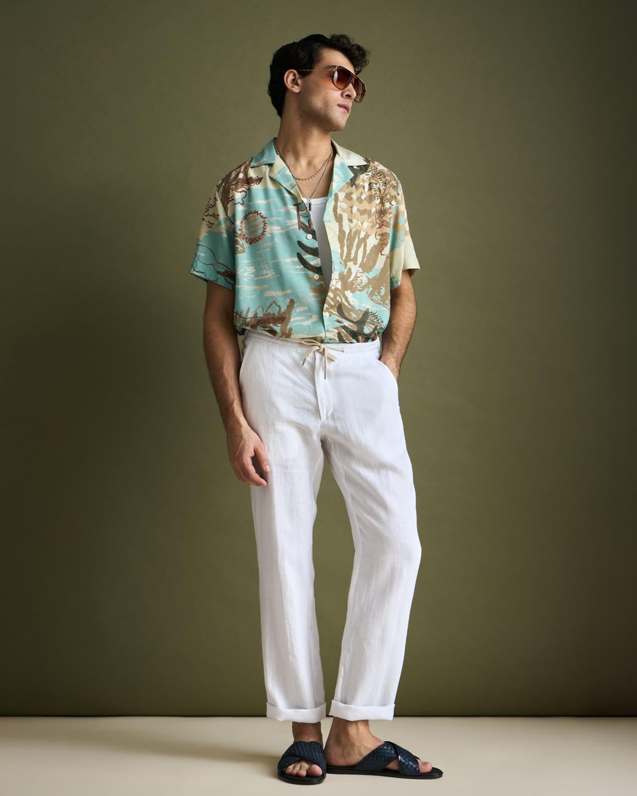 Un mannequin porte une chemise à manches courtes à imprimé tropical avec un pantalon blanc à cordon de serrage, des sandales noires à enfiler, et accessoirise avec des colliers et des lunettes de soleil