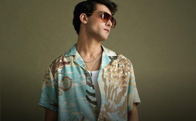 A fashion model wears a short-sleeved tropical print shirt with white drawstring trousers, black slide sandals, and accessorizes with necklaces and sunglass