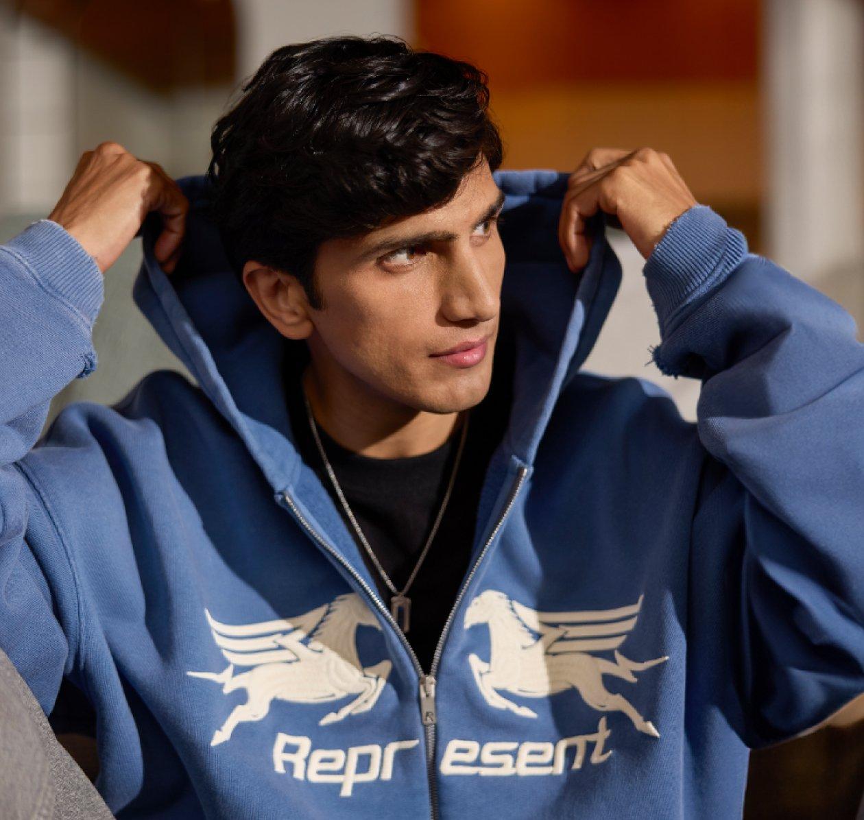 Un homme portant un hoodie bleu de la marque Represent avec un t-shirt noir et un jean gris