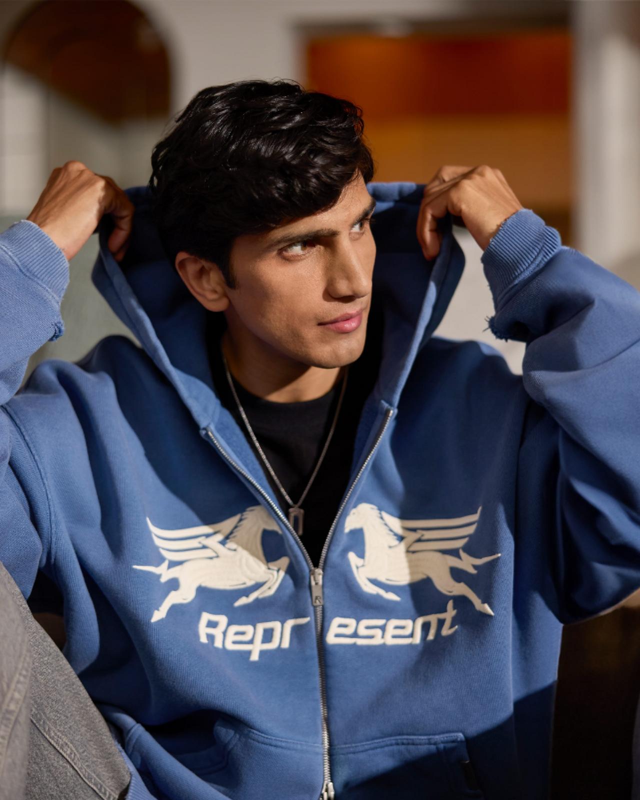 A man wearing a blue Represent designer hoodie with black t-shirt and grey jeans