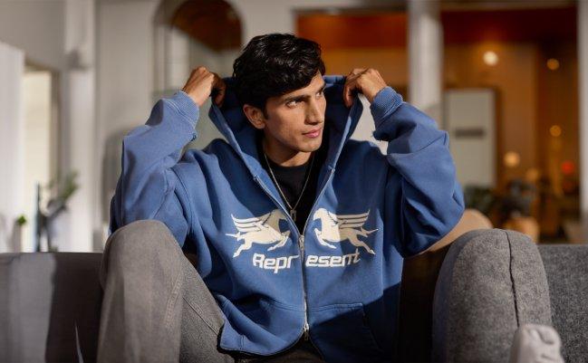 Un homme portant un hoodie bleu de la marque Represent avec un t-shirt noir et un jean gris