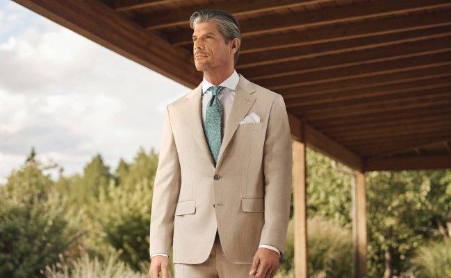 Un homme portant un complet beige clair avec une chemise blanche, une pochette de costume et une cravate bleu pâle