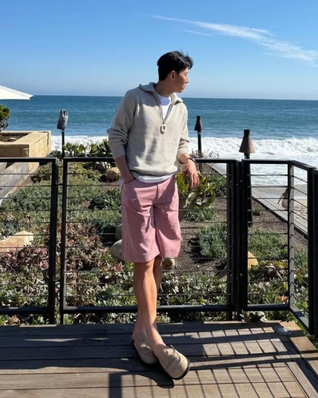 Son Heung standing on the deck at the beach side wearing quater-zip sweater with pink shorts and soft mules