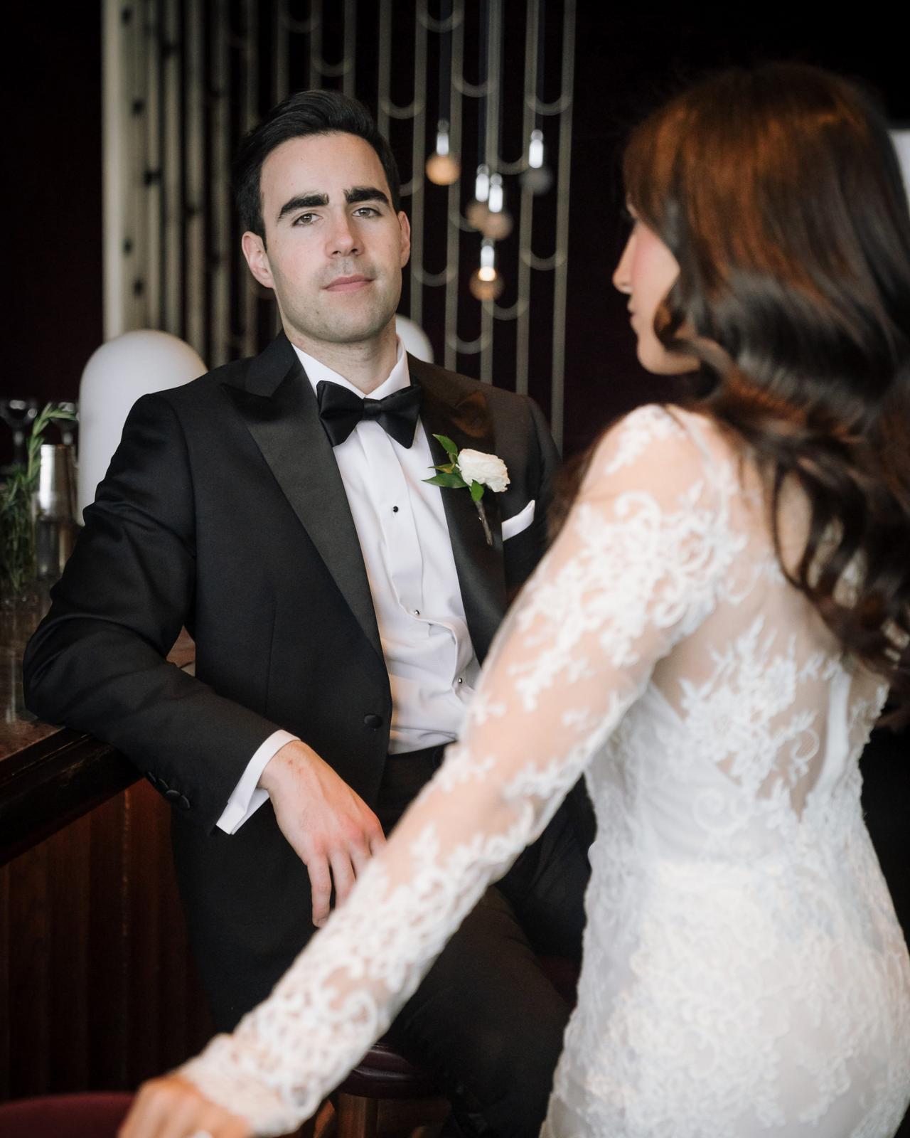 A bride in a long-sleeved lace wedding gown and a groom in a black tuxedo with a white rose boutonniere