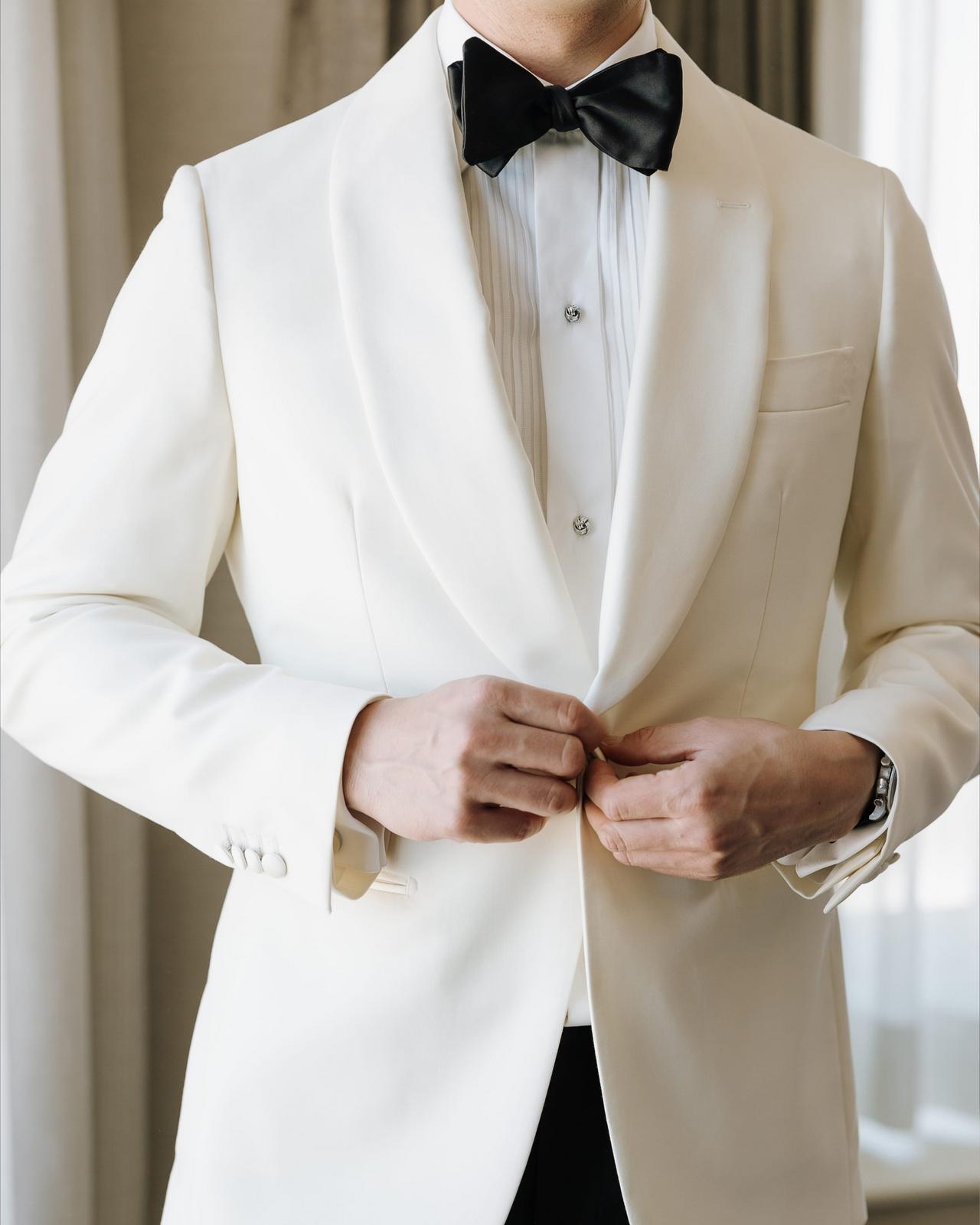 A groom wearing an off-white tuxedo with white shirt and black bow tie