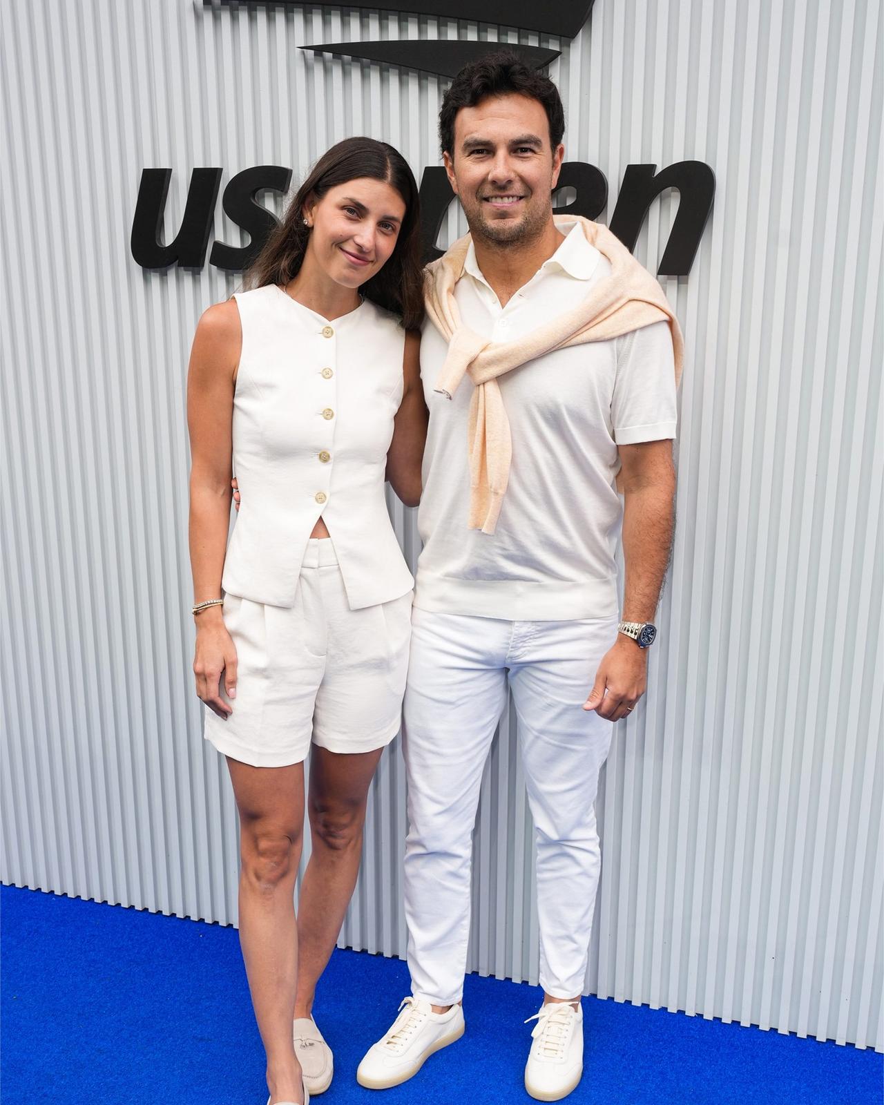 Checo Perez wearing a white polo with slim fit pants and sweater on his shoulder standing with his wife