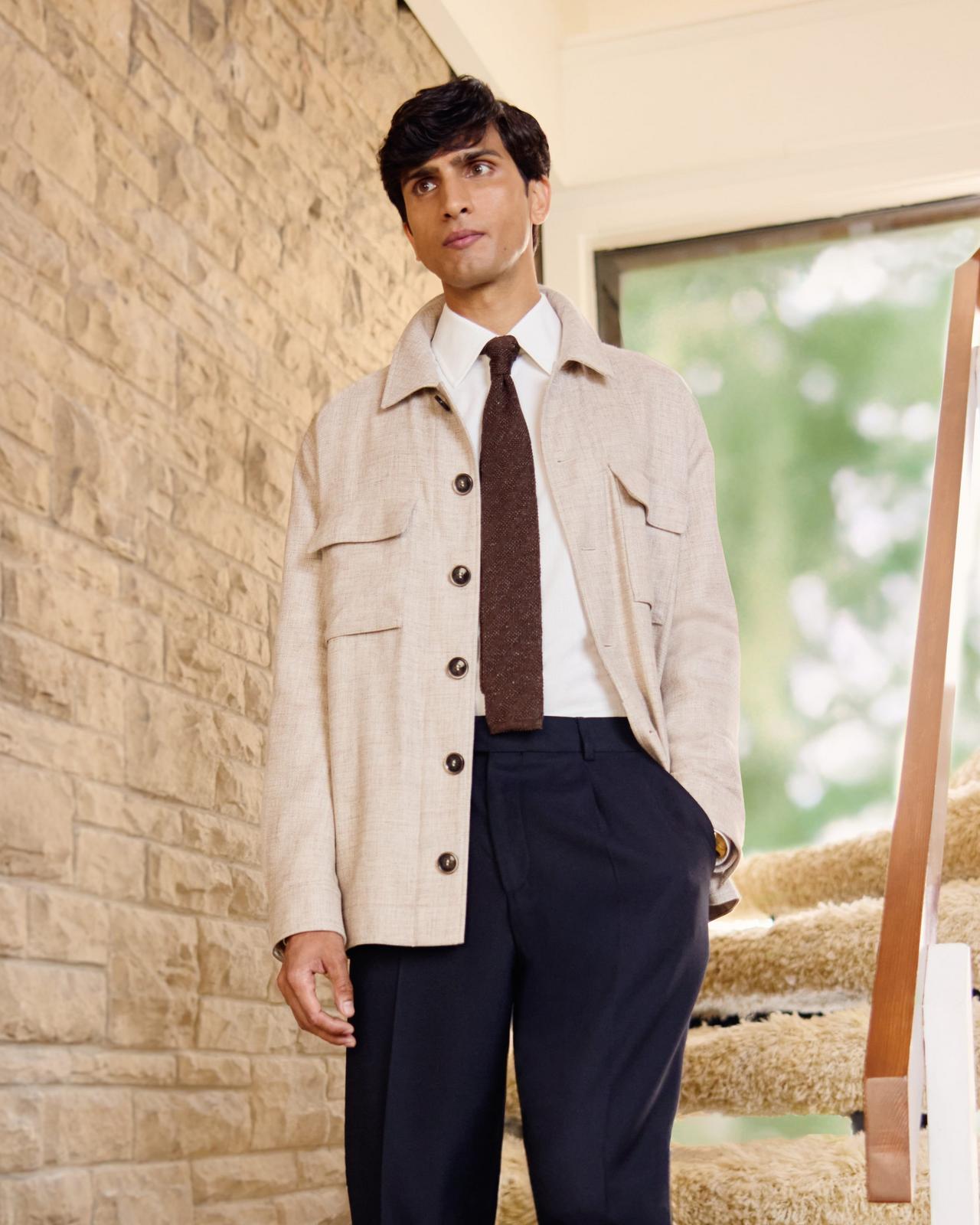 A male model wearing a tan colored coat, white dress shirt, navy trousers and a brown tie