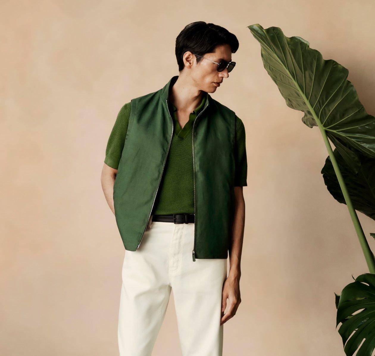 Mannequin de mode portant un gilet zippé vert foncé par-dessus une chemise verte texturée à manches courtes, assorti à un pantalon blanc et une ceinture noire