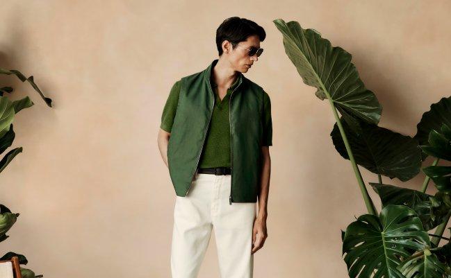 Fashion model wearing a dark green zip-up vest over a textured green short-sleeve shirt, paired with white trousers and a black belt