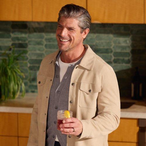 A man wearing a light cream jacket with grey sweater and white t-shirt