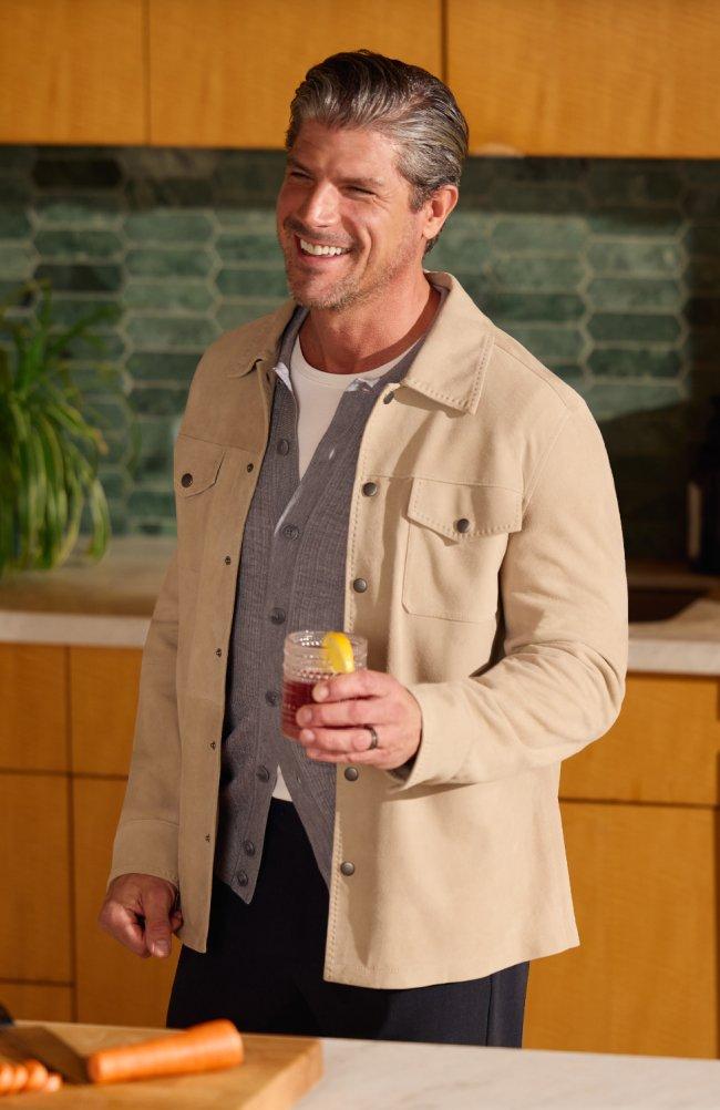 A man wearing a light cream jacket with grey sweater and white t-shirt