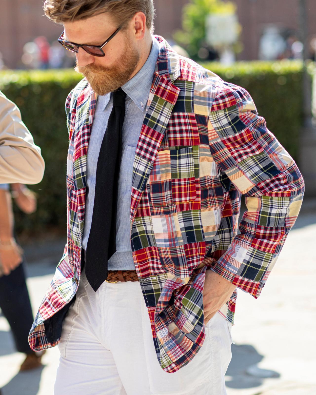 A man wearing a colorful patchwork plaid blazer over a light blue dress shirt and black tie, paired with white trousers
