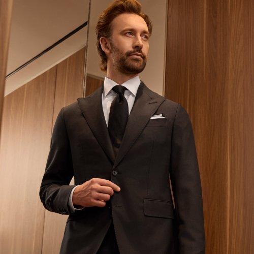 Un homme portant un manteau noir, une chemise blanche et une cravate noire se tenant près du miroir