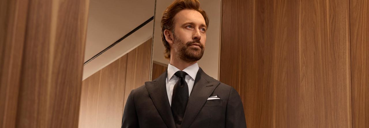 A man wearing a black coat, white shirt and black tie standing near the mirror