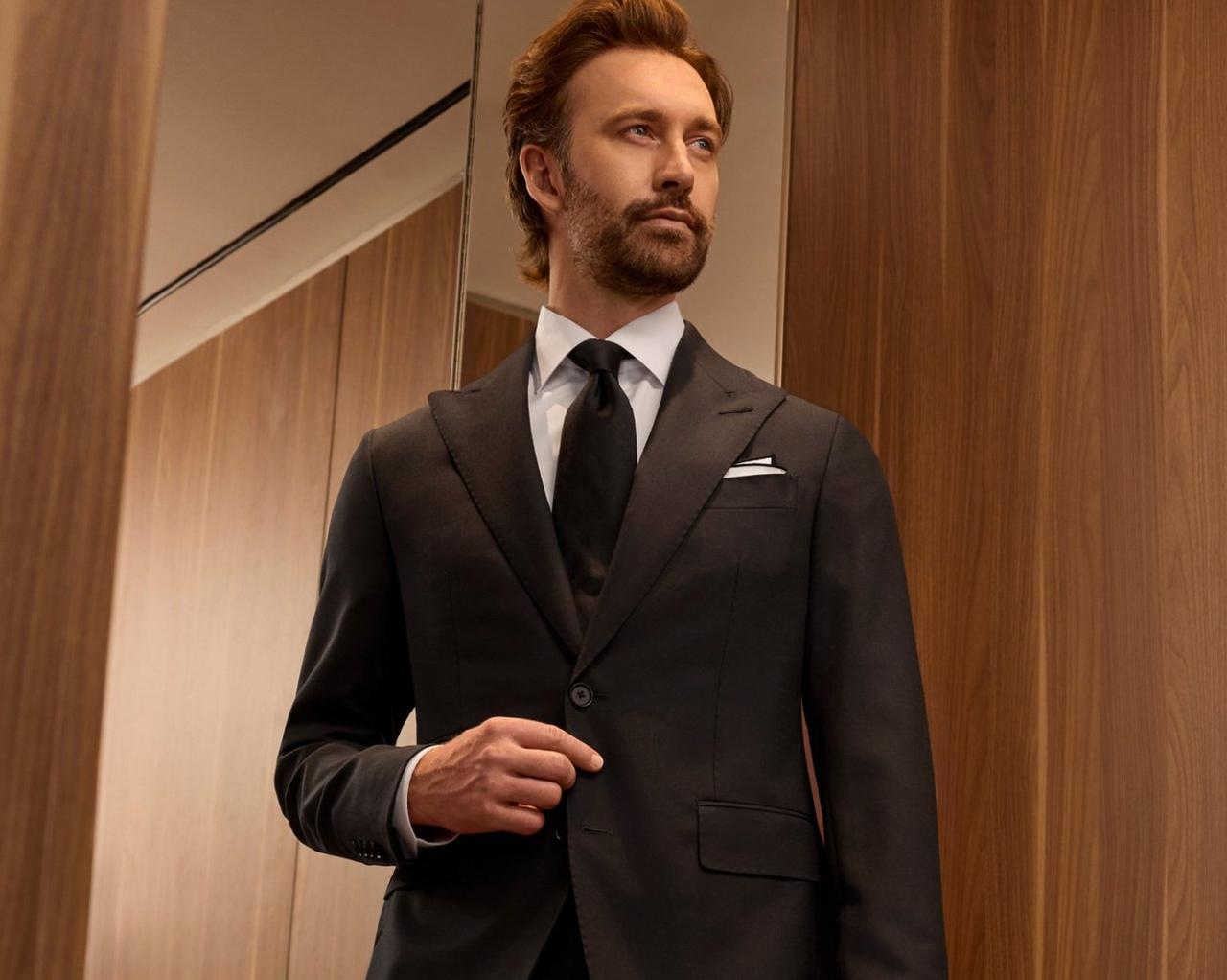A man wearing a black coat, white shirt and black tie standing near the mirror