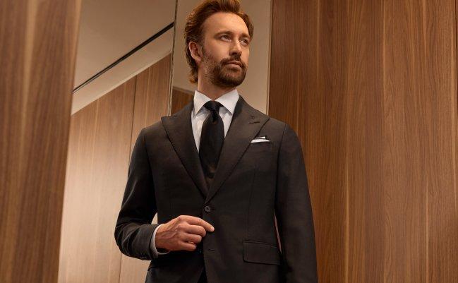 Un homme portant un manteau noir, une chemise blanche et une cravate noire se tenant près du miroir