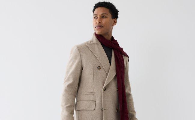 A male model wearing a tan coat styled with a dark grey sweater, red scarf and tan gloves