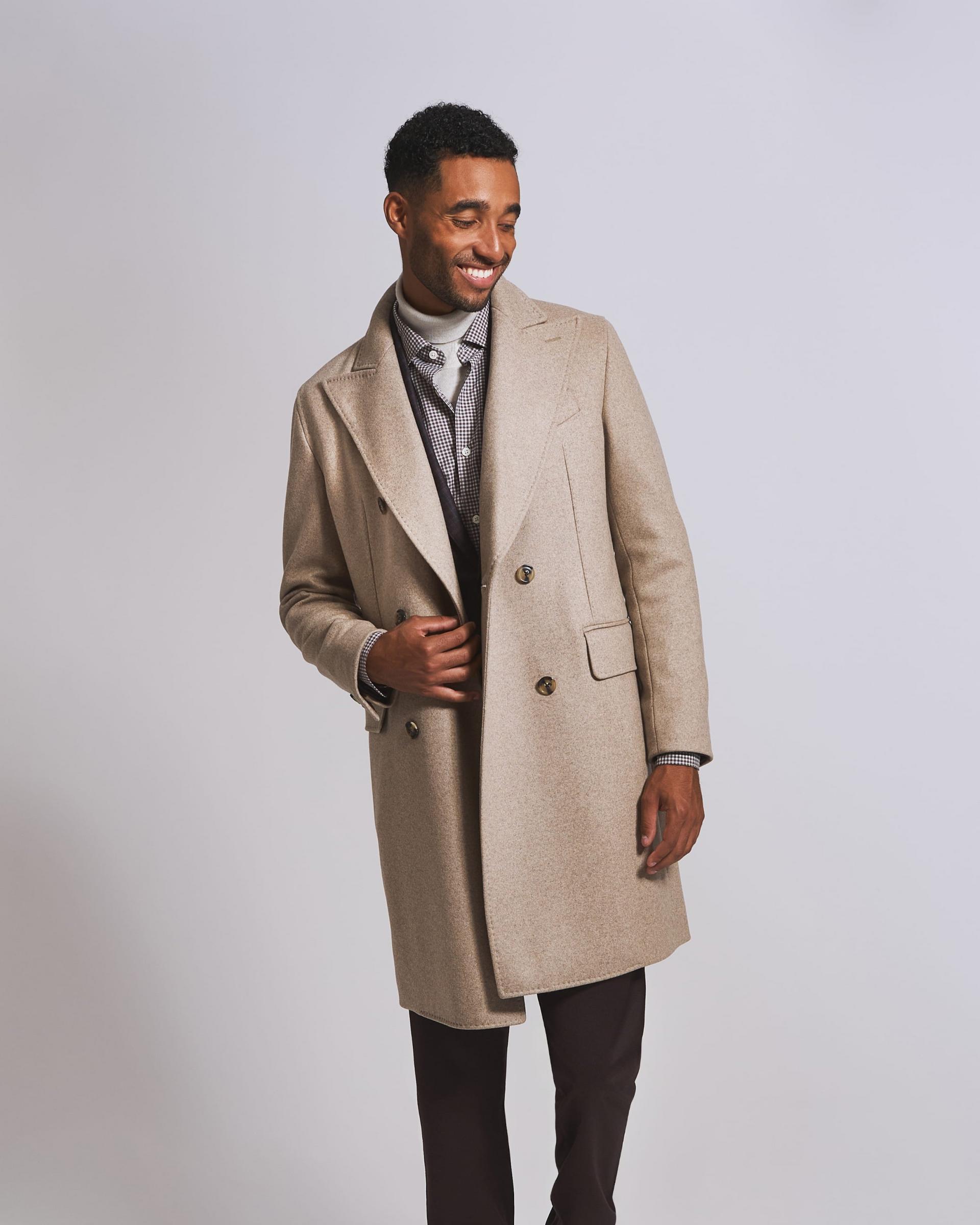 A male model wearing a long tan colored overcoat styled with a white turtleneck sweater
