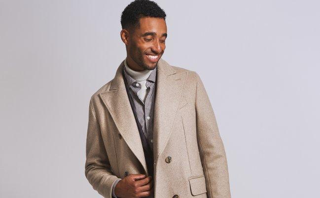 A male model wearing a long tan colored overcoat styled with a white turtleneck sweater