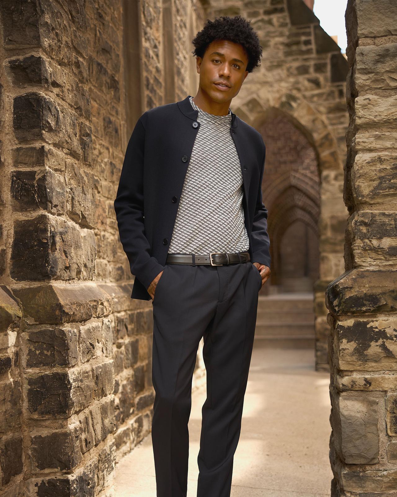 Homme portant une veste noire boutonnée par-dessus un chandail gris à motifs, avec un pantalon et une ceinture noirs, debout devant une arche en pierre